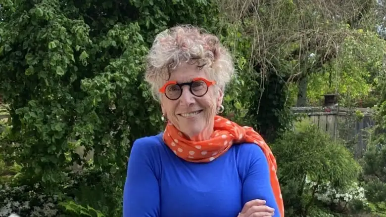 A woman wearing orange and blue circle glasses with short curly grey hair. 