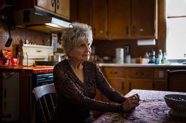 Alice Munro, with short gray hair and wearing a brown blouse, sits at a table in a modest kitchen. Her hands are clasped in front of her on the colorful tablecloth.