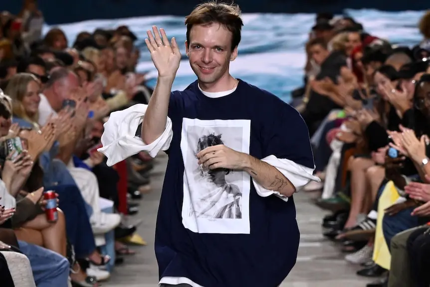 A man in pants and a blue t-shirt with a bell-sleeve shirt underneath waves, running down an aisle to people clapping.
