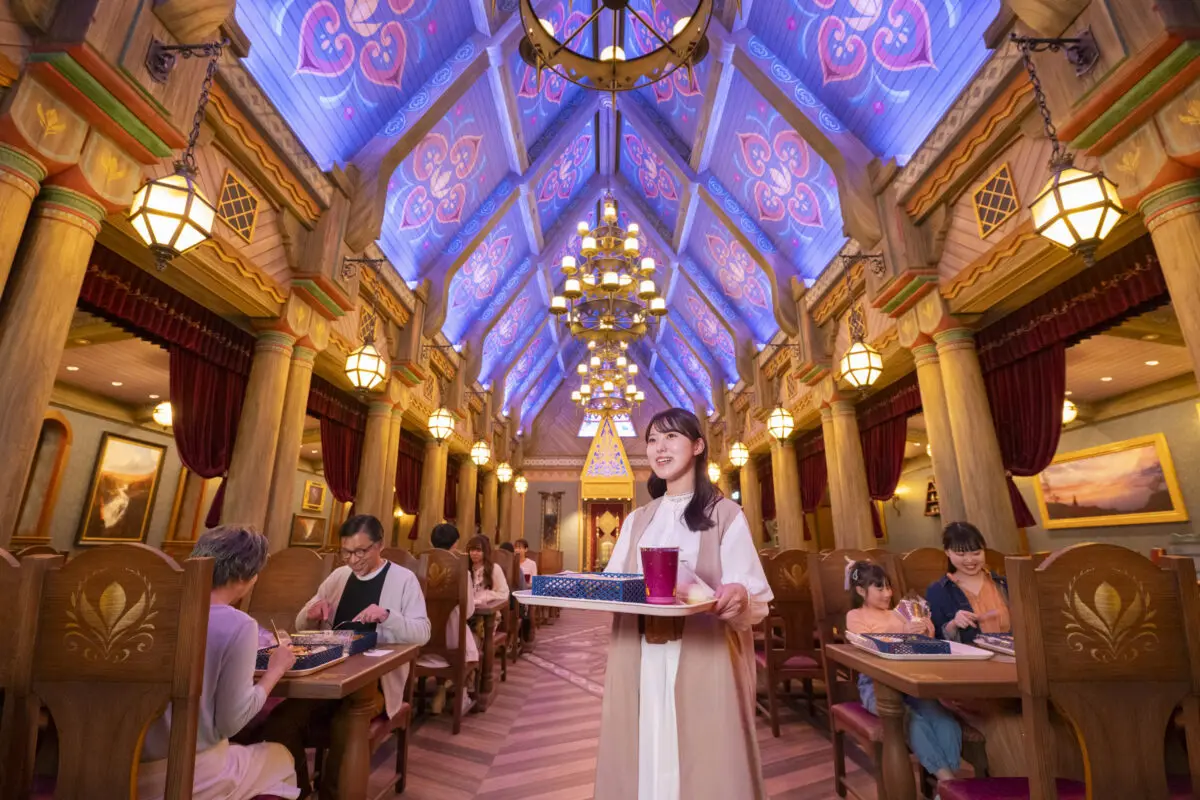 A waitress serving drinks in a vibrant, ornately decorated restaurant with high ceilings, chandeliers, and patrons dining at wooden tables.