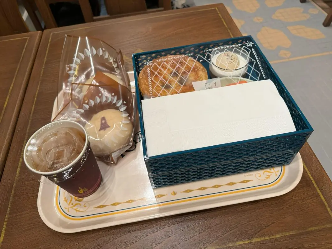 A tray holds a cup of iced coffee, several wrapped pastries, and a basket containing a pastry with lattice crust, small containers, and a napkin.