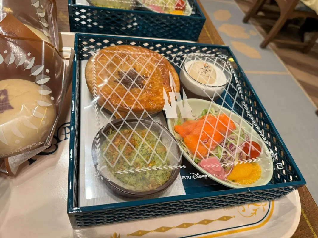 A neatly arranged bento box containing a pastry, a bowl of rice with toppings, a small salad with orange slices, and a dessert in a small bowl, placed on a tray with a partially visible wrapping nearby.