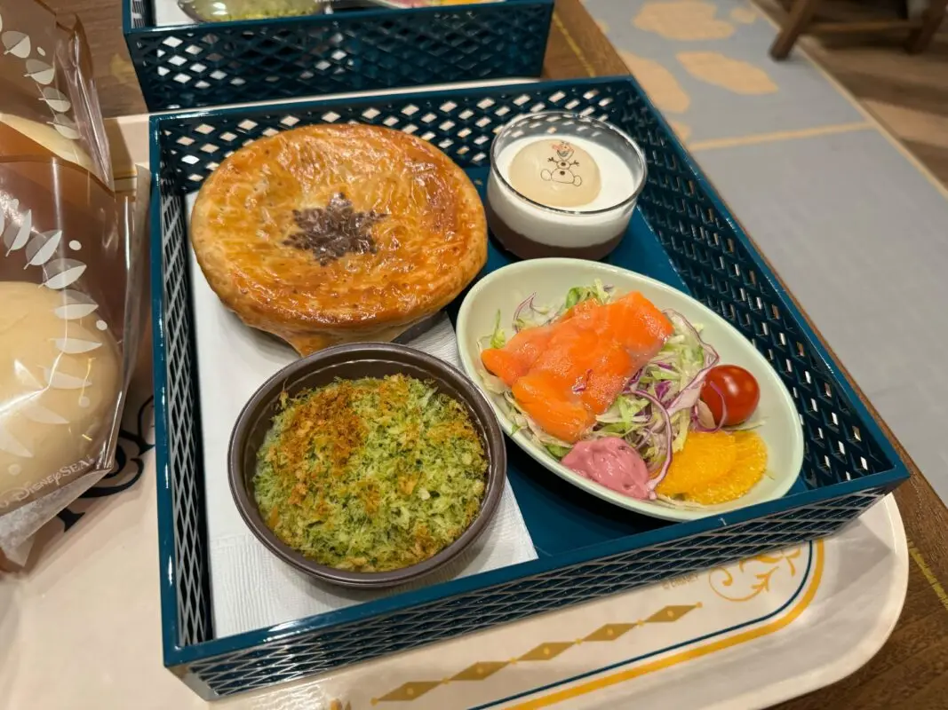 A tray with various dishes: a baked pastry, a bowl of leafy greens topped with breadcrumbs, a salad with smoked salmon, tomatoes, and orange slices, and a small container of creamy dressing.