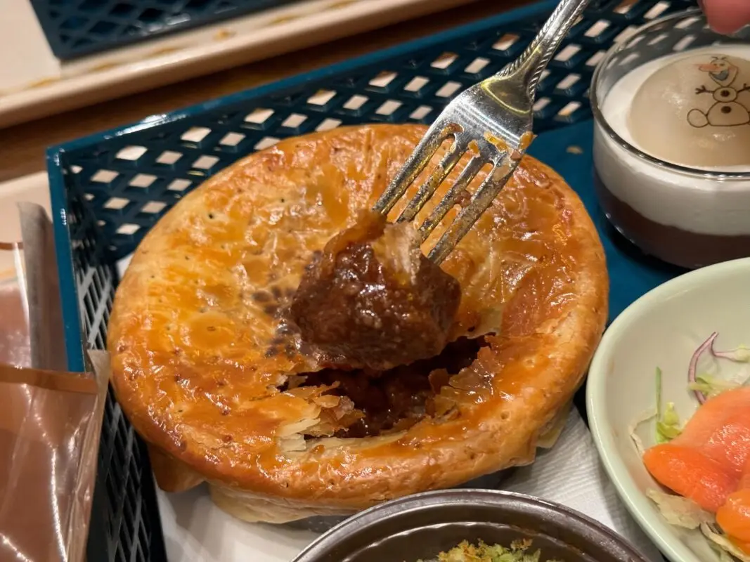 A fork lifts a piece of meat from a golden-brown pie placed on a tray beside a bowl of salad and a cup of dessert.