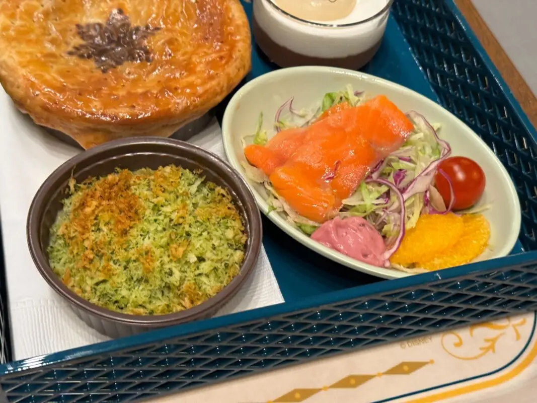 A tray containing a baked dish with a pastry crust, a small bowl of gratin, and a side salad with smoked salmon, cherry tomatoes, and a purple dressing.