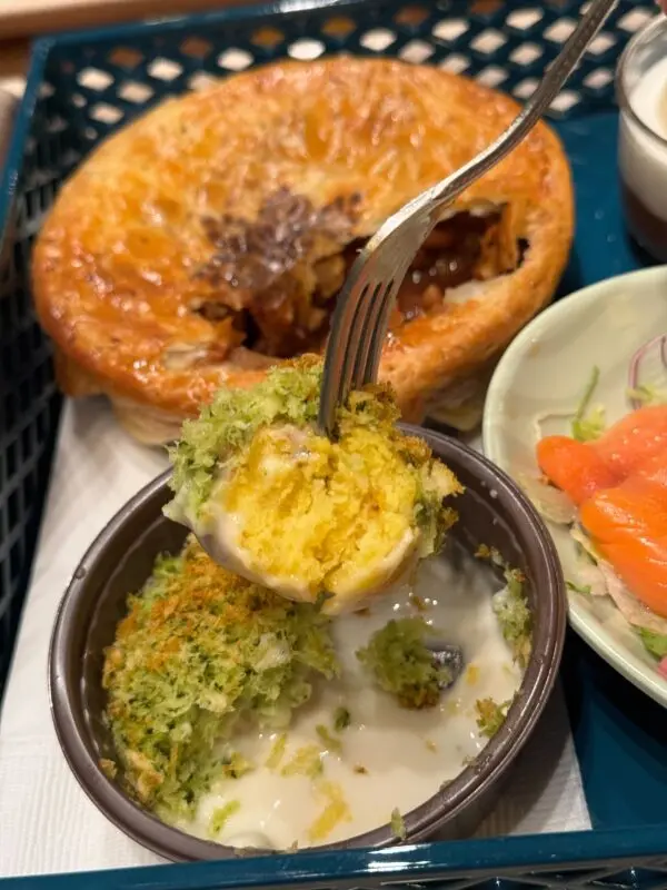 A fork holding a piece of bread pudding with cream sauce, in front of a small bowl. In the background, there is a pie and a plate with salmon and vegetables.