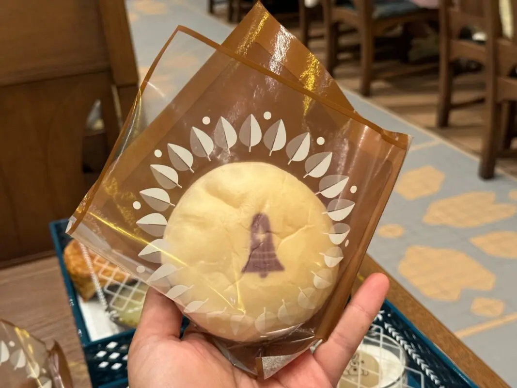 A hand holds a wrapped pastry with a tree design printed on the packaging. The background shows a wooden table and chairs.