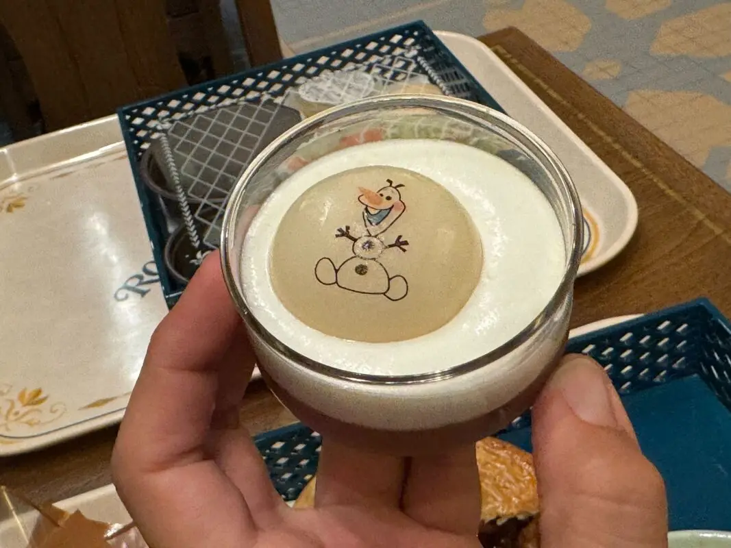 A hand holding a dessert topped with whipped cream and a printed image of a snowman character. Other food items and trays can be seen in the background.