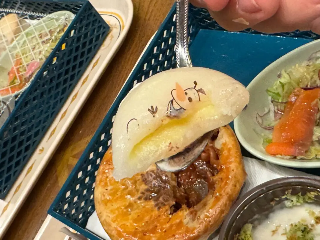 A spoon holding a steamed bun with a printed cartoon character, covering a meat-filled pastry, next to a plate of vegetables and salad.