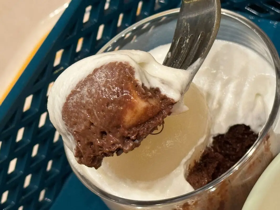 A close-up of a spoonful of chocolate mousse topped with whipped cream, taken from a glass container on a blue plastic tray.