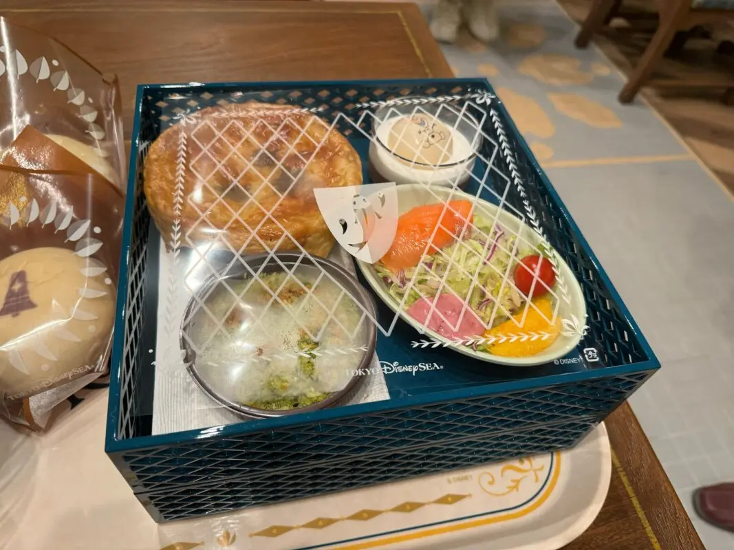 A neatly packed food tray includes a pie, soup, salad, and a dessert, enclosed in a blue lattice-patterned container placed on a light-colored tray.