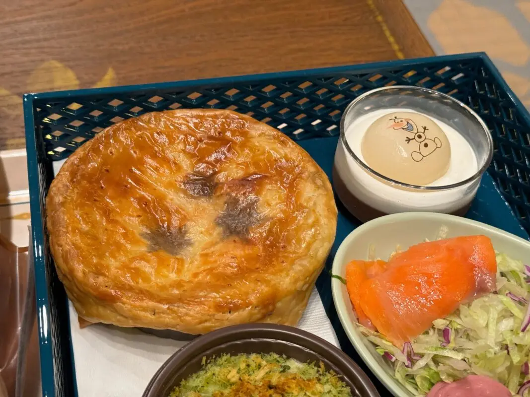 A tray with a baked pie, a small bowl of white custard dessert with a cartoon character, a green bowl with salmon and mixed salad, and a small dish of breadcrumb-topped food.