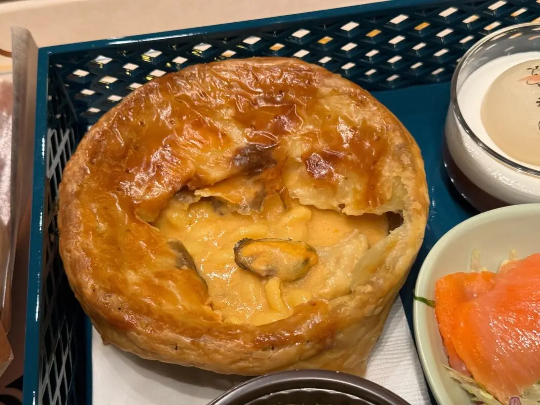 Photo of a golden-brown puff pastry pie with a creamy filling, accompanied by a small dish of what appears to be smoked salmon, on a blue tray.