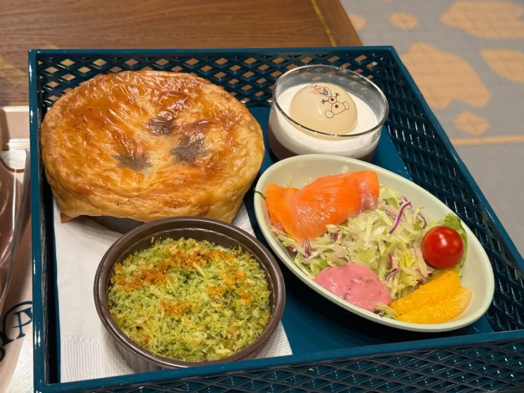 A meal tray featuring a baked pie, a small bowl of green gratin, a salad with lettuce, salmon, a cherry tomato, and an orange slice, and a dessert served in a small glass with a cartoon design.