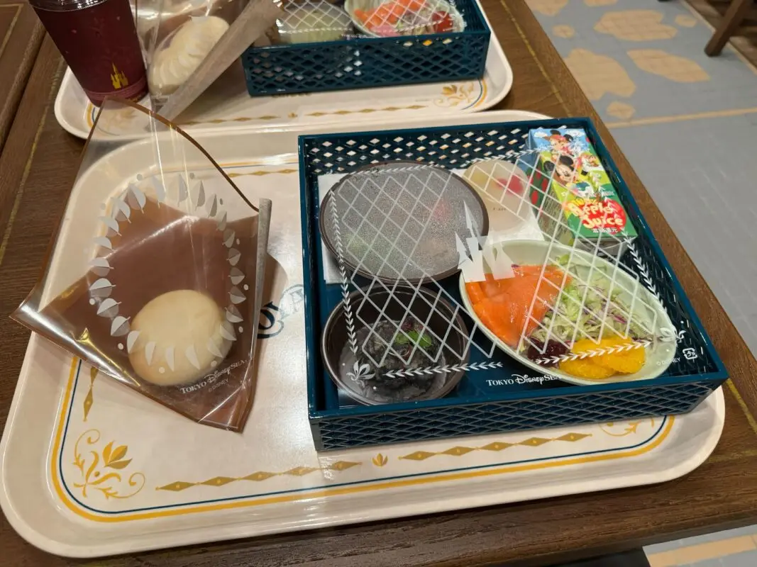 A tray of food featuring a steamed bun wrapped in plastic, and a bento box with rice, vegetables, a slice of orange, and a piece of dessert.