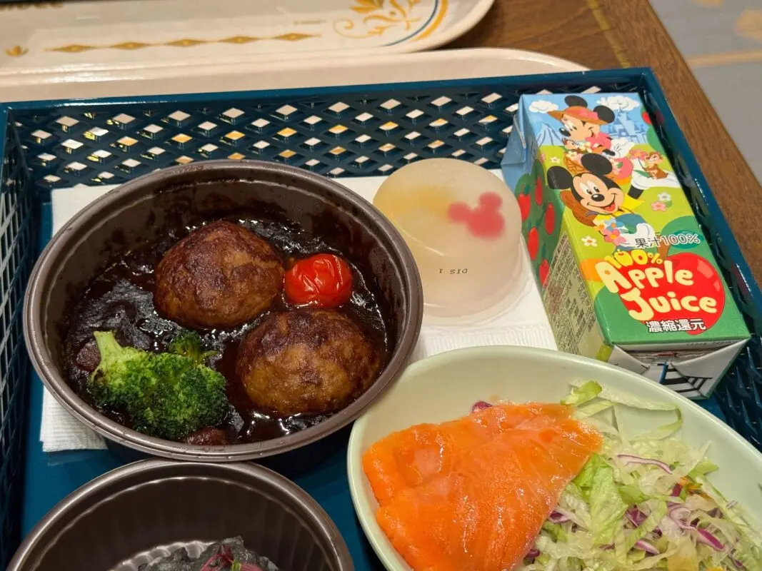 A tray with a meal including two meatballs in a brown sauce with broccoli and a cherry tomato, a small fruit jelly, smoked salmon with salad, and a box of 100% apple juice.