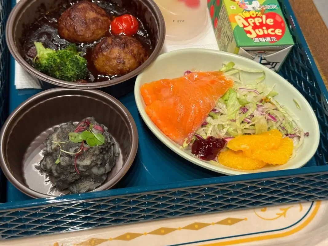 A meal tray with meatballs, broccoli, and cherry tomato, a side of mashed potatoes, salad with smoked salmon, and citrus slices, accompanied by a carton of apple juice.