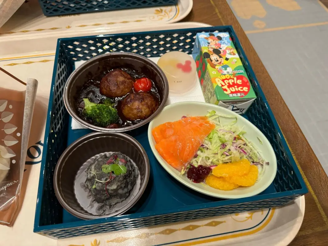 A tray with meatballs in sauce, a small dessert, salmon with salad and orange slices, black bread with herbs, and a carton of apple juice.