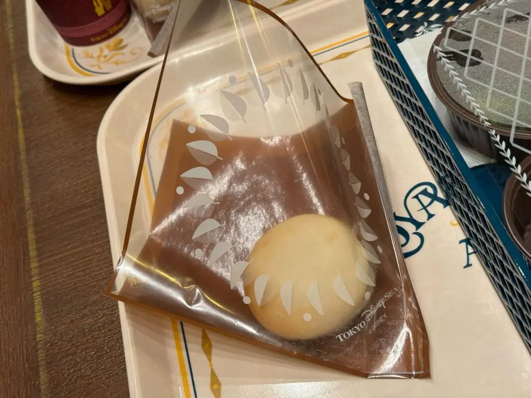 A single round pastry wrapped in a transparent plastic casing with leaf designs on a beige tray.