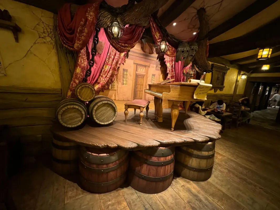 A small stage with barrels as the floor, featuring a wooden desk and chair, against a backdrop of red curtains and a painted village scene.