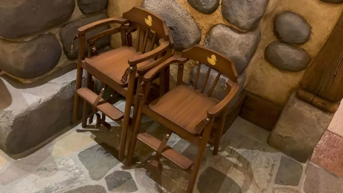 Two wooden high chairs for children are placed side by side against a stone wall. The chairs feature armrests and a small footrest.