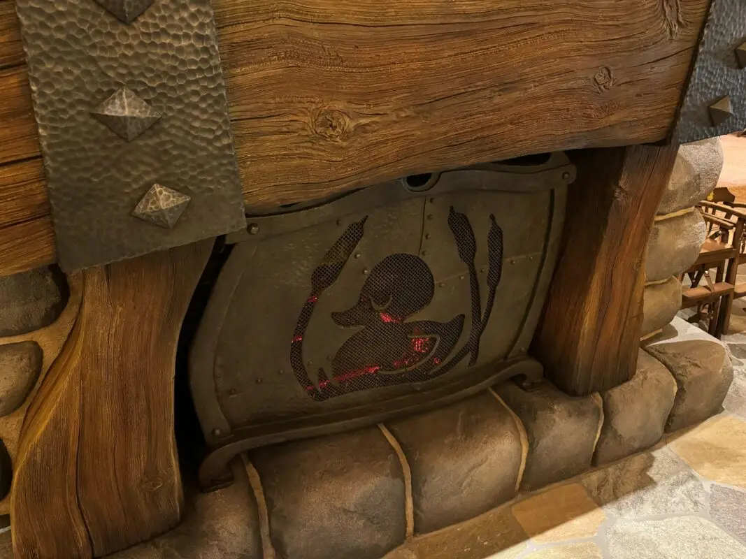 A rustic wooden fireplace with a metal grate displaying glowing red embers inside, surrounded by stone and wooden details.