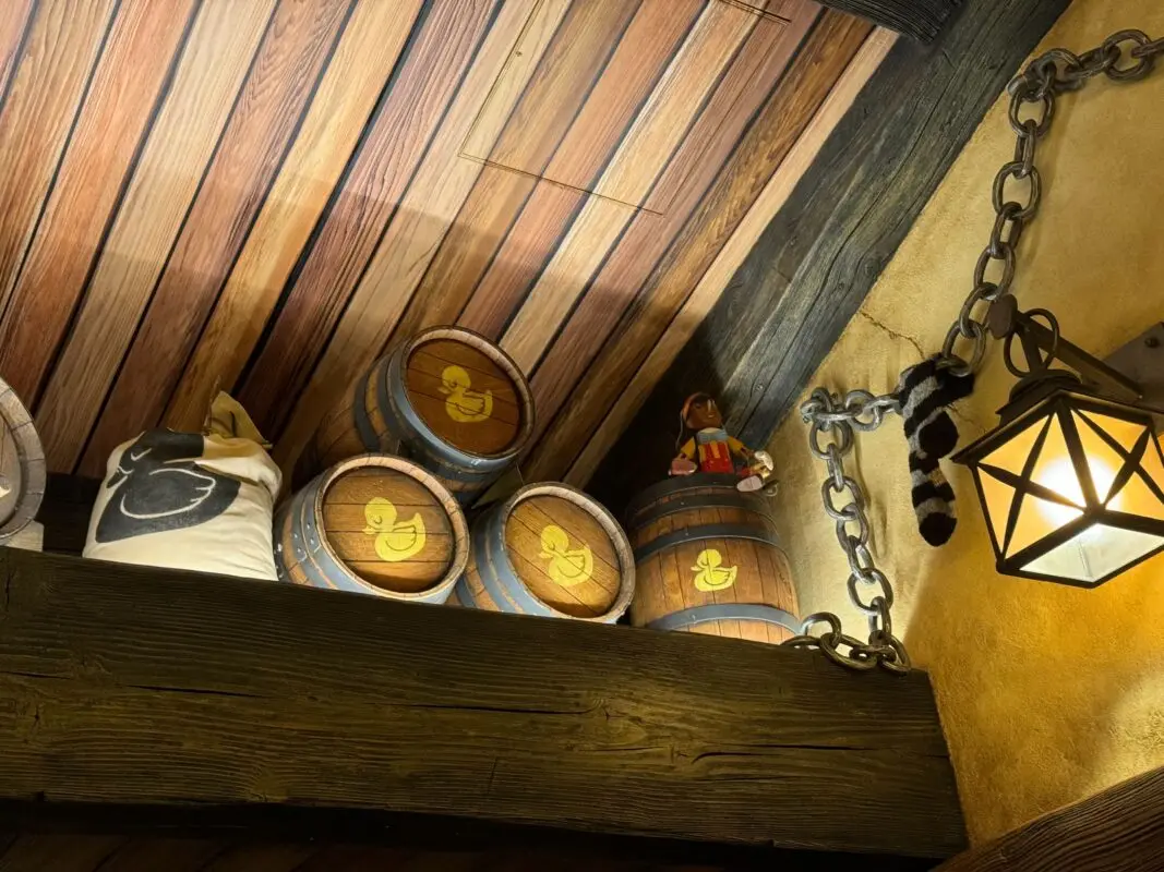 Barrels with yellow duck symbols, a stuffed toy, and a sack are stored on a wooden shelf below a wood-paneled ceiling. An iron chain and a lantern are attached to the wall on the right.