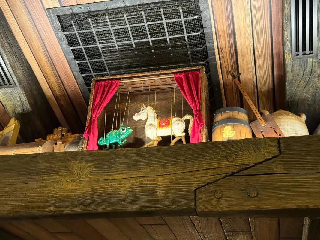 Wooden shelf with a small puppet theater scene featuring a horse and a chameleon on strings. The background includes a barrel, pottery, and wooden instruments. The setting includes wooden walls.