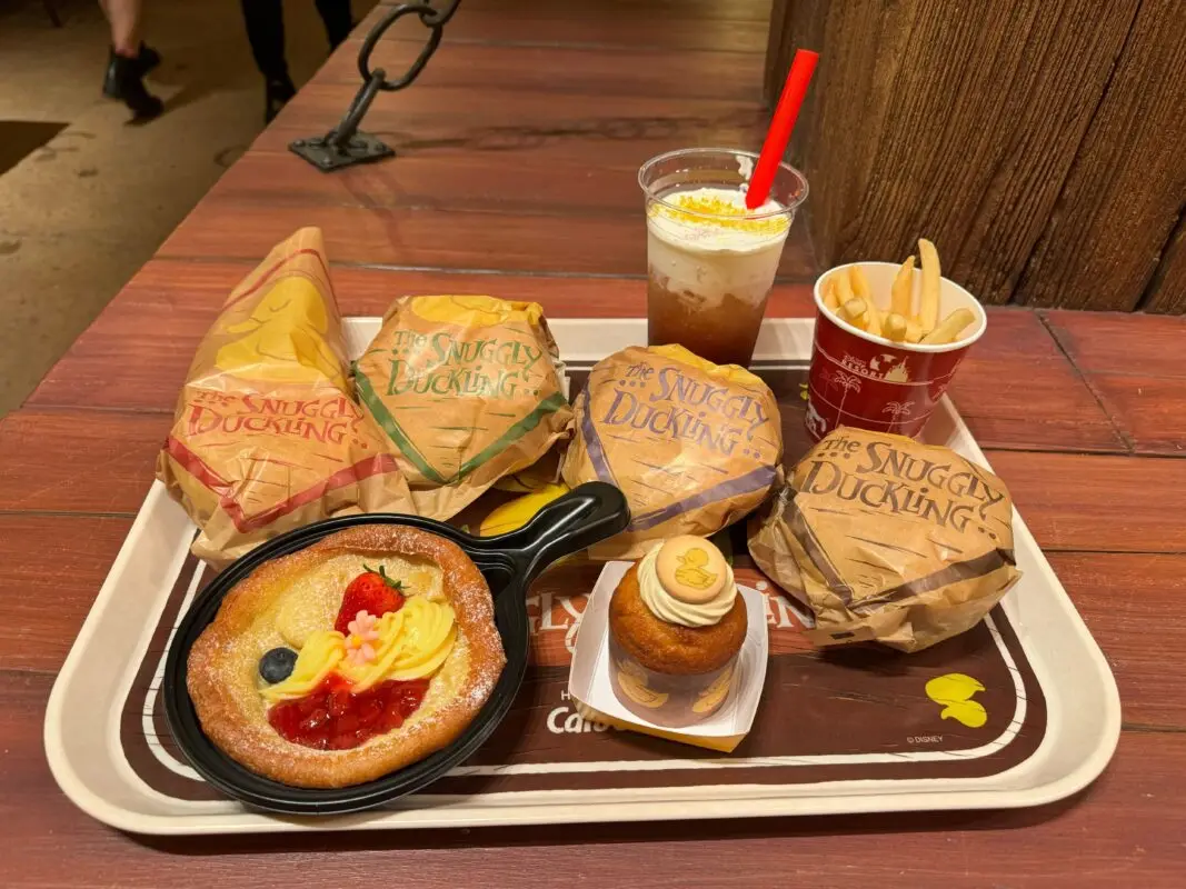 A tray holds five wrapped sandwiches, a small cup of fries, a frosted beverage with a red straw, a fruit-topped dessert in a skillet, and a frosted cupcake on a wooden table.