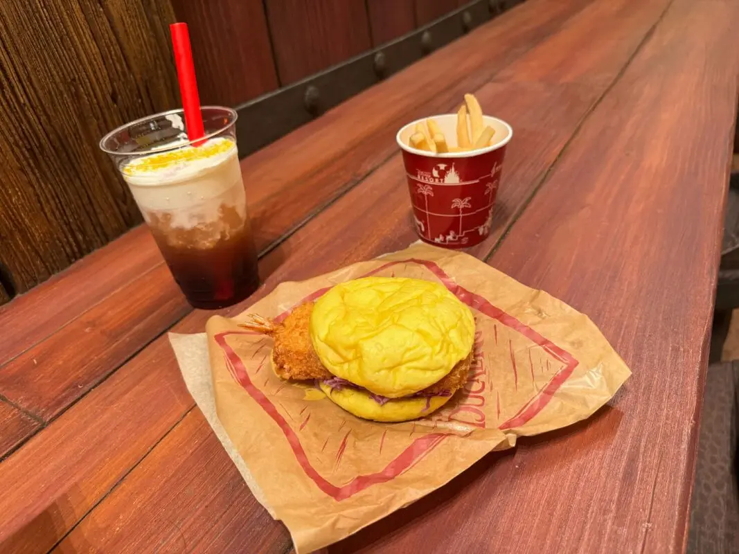 A chicken sandwich on a paper wrapper, a cup of french fries, and a cold drink are on a wooden table.