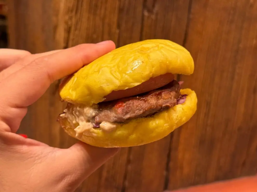 A hand holding a cheeseburger with a yellow bun, beef patty, tomato slice, mayo, and ketchup against a wooden background.