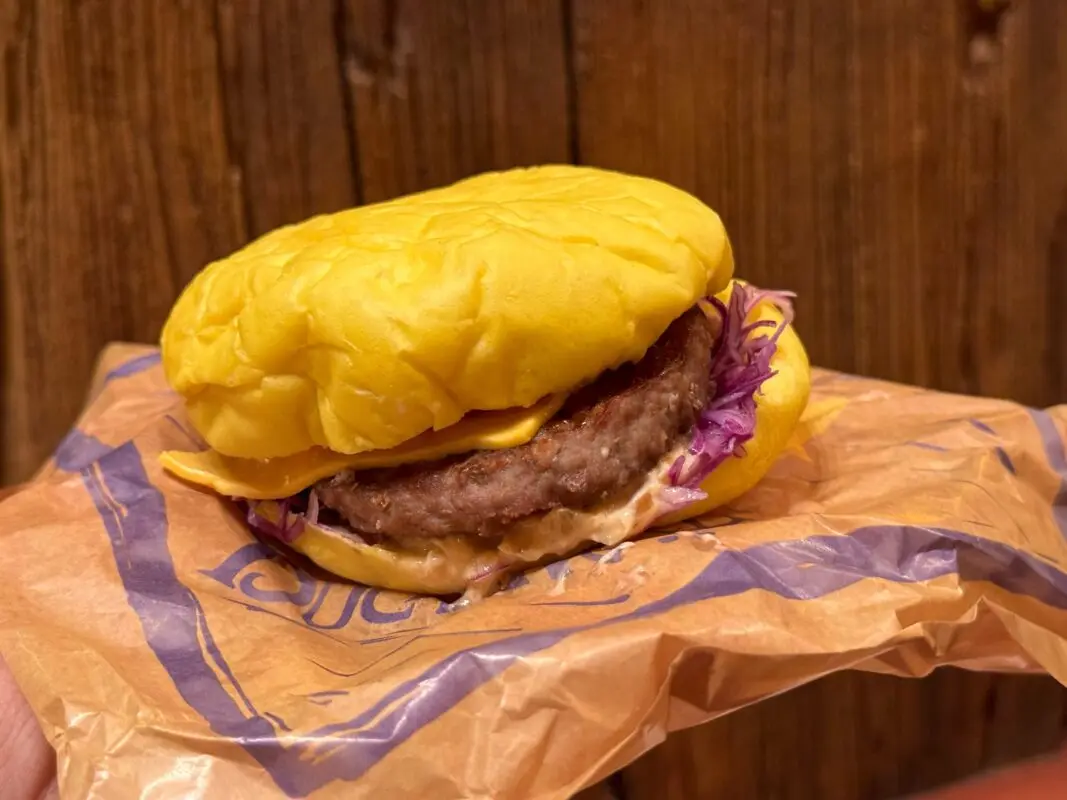 A close-up of a cheeseburger with a single patty, cheese, shredded lettuce, and sauce, wrapped in a brown paper wrapper.