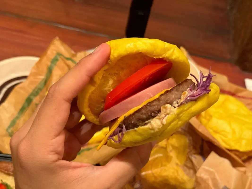 A close-up of a hand holding a burger with a yellow bun, beef patty, slice of processed meat, tomato, purple cabbage, and sauce.