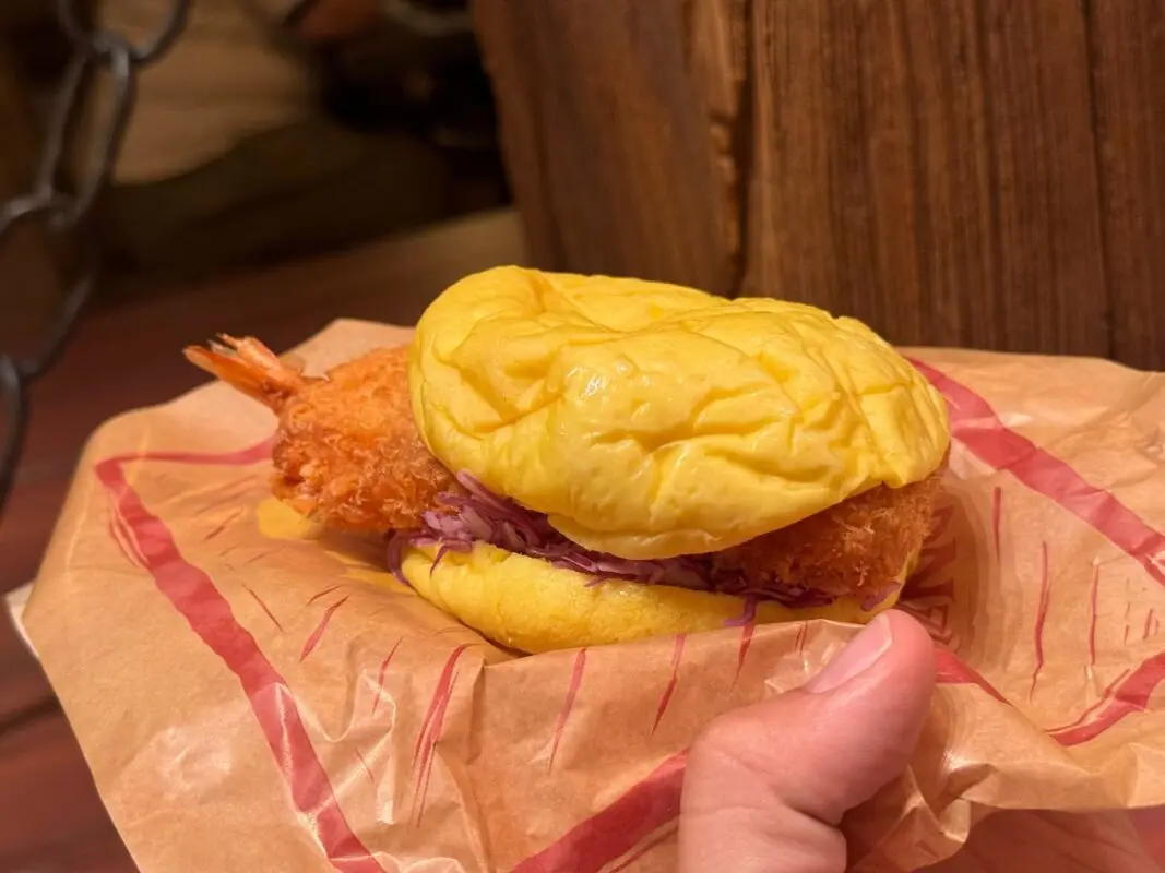 A person holds a sandwich with a fried shrimp patty and purple coleslaw in a yellow bun, wrapped in brown paper.