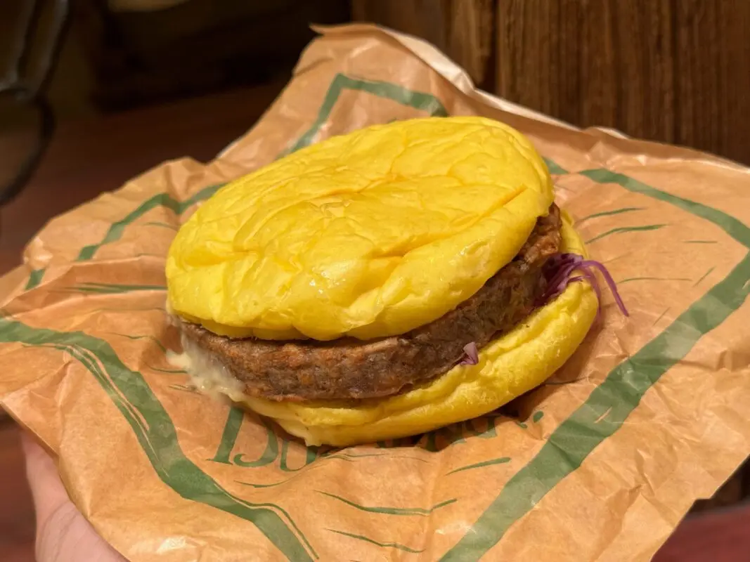 A cooked burger with a yellow bun, meat patty, and a small amount of toppings held in a brown paper wrapper.