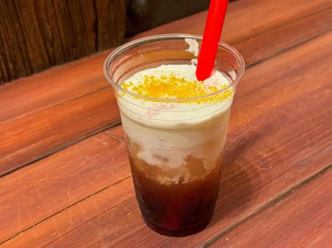 A plastic cup with a layered cold beverage, featuring dark liquid on the bottom, white foam on top, and yellow granules sprinkled over the foam, with a red straw inserted. Placed on a wooden surface.