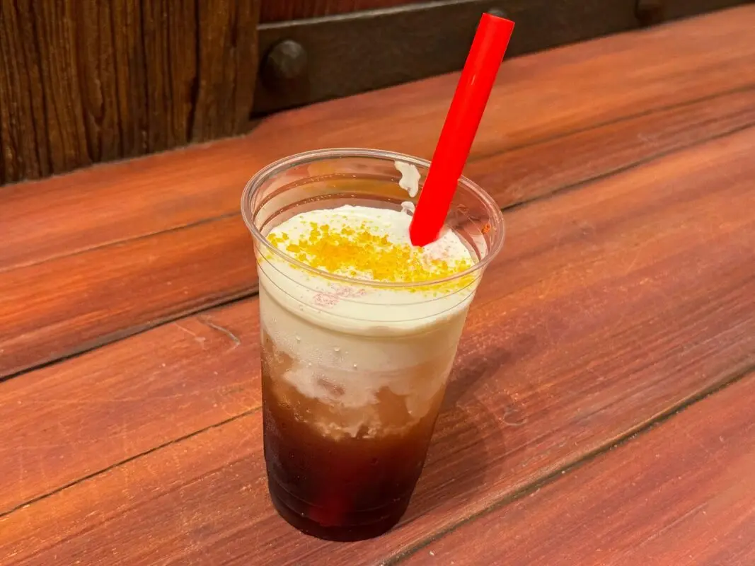 A plastic cup filled with a frothy, layered beverage garnished with yellow crumbs and a red straw, placed on a wooden surface.