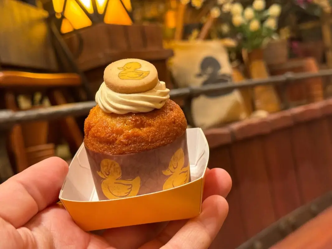 REVIEW: “I’ve Got a Nightmare” - The Snuggly Duckling Restaurant Disappoints in Tangled Kingdom at Fantasy Springs 16 A hand holding a small cupcake with creamy frosting and a duck-shaped cookie on top, set in a decorative wrapper with a duck motif. The background shows a warmly lit interior with various decor items.
