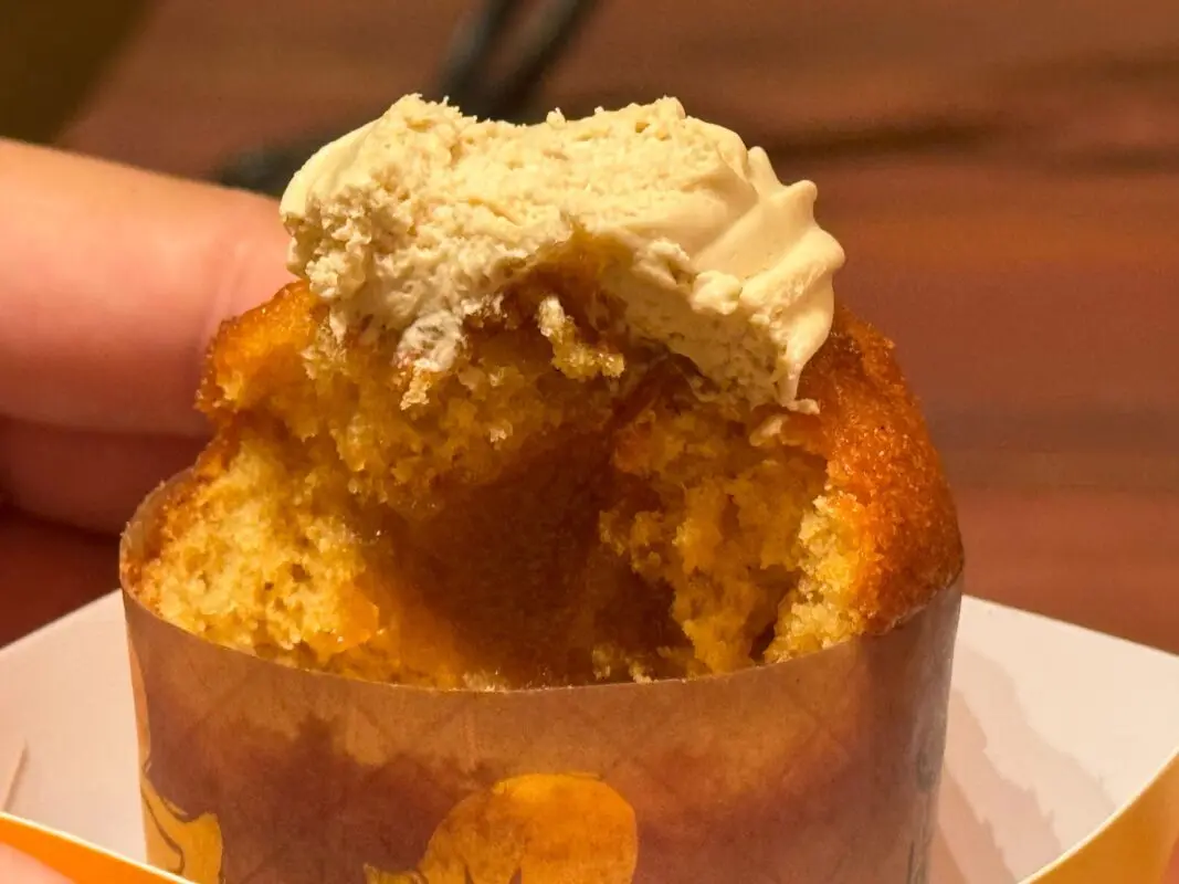 Close-up of a half-eaten cupcake with light brown frosting on top, being held in a hand over an orange patterned paper wrapper.