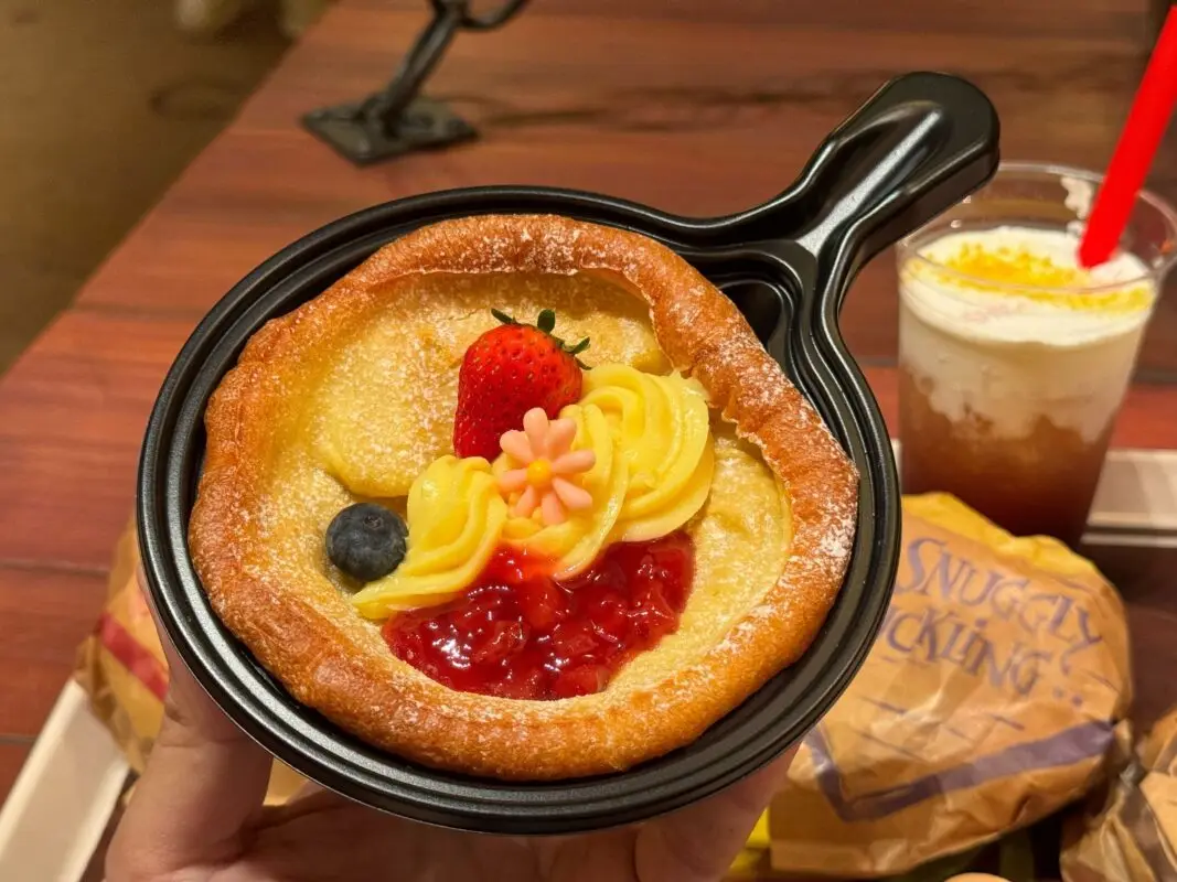 A small cake with custard, strawberry, blueberry, and flower-shaped decorations in a black dish alongside a drink and food items on a wooden table.