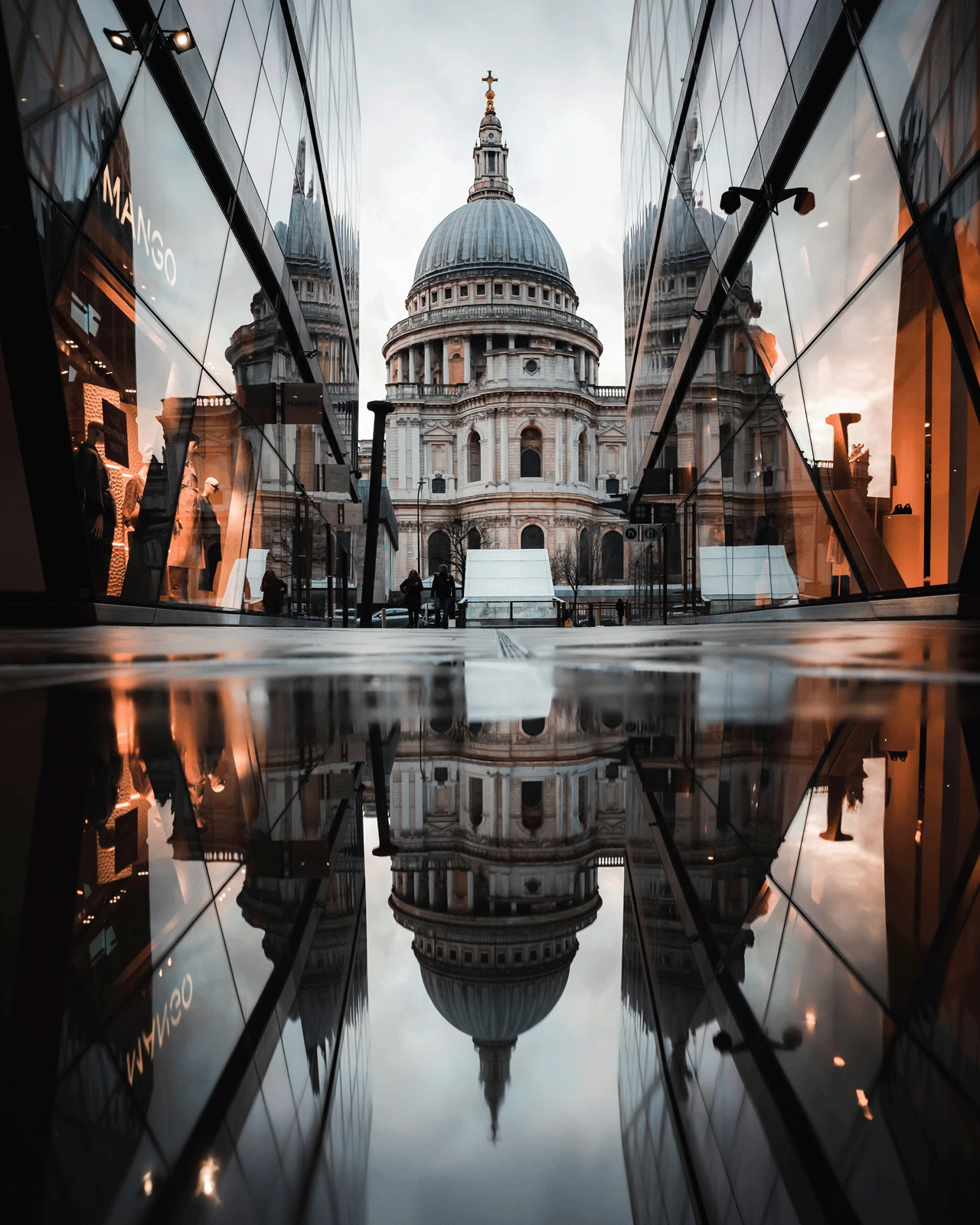 A view of St. Paul's Cathedral in London, framed by modern glass buildings on either side, with a perfect reflection of the scene on a wet surface in the foreground. The sky is overcast, adding a dramatic atmosphere to the image.