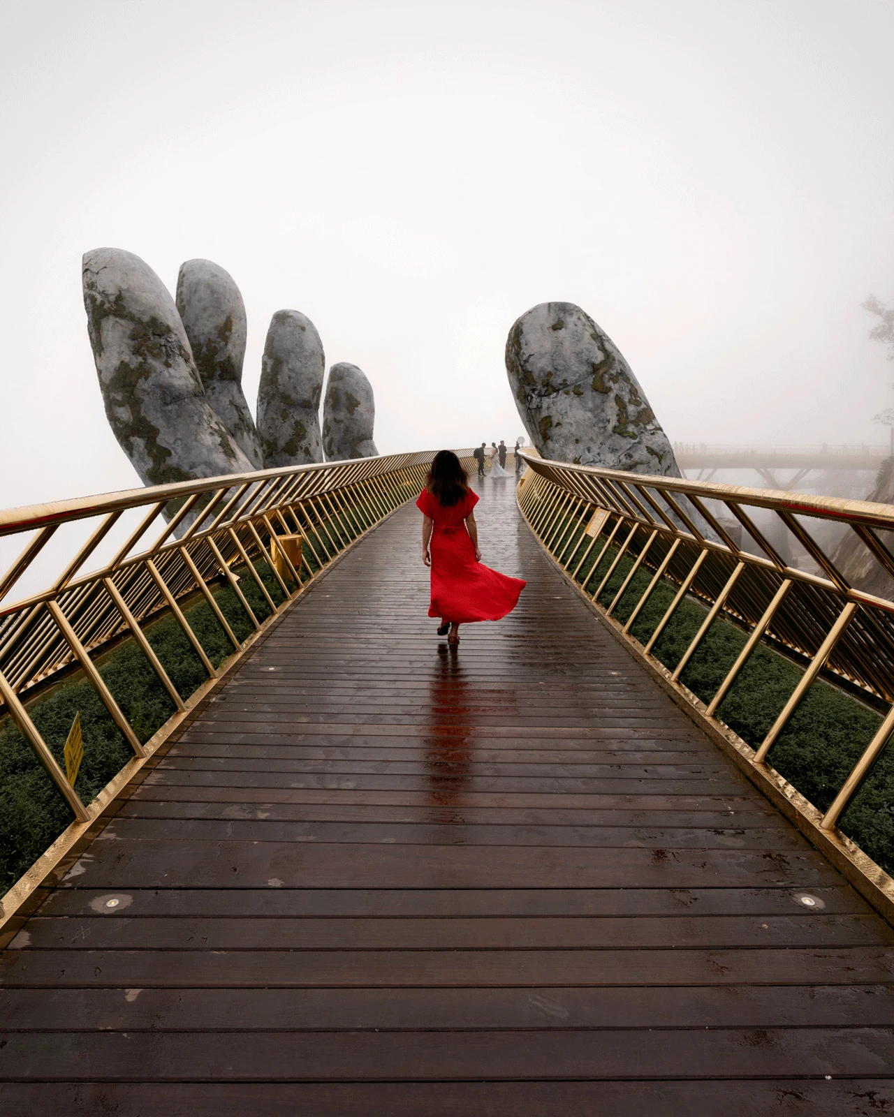 A woman in a flowing red dress walks across the Golden Bridge in Da Nang, Vietnam. The bridge is held up by two giant stone hands and surrounded by mist, creating a mystical atmosphere. The bridge has golden railings and a wooden path.
