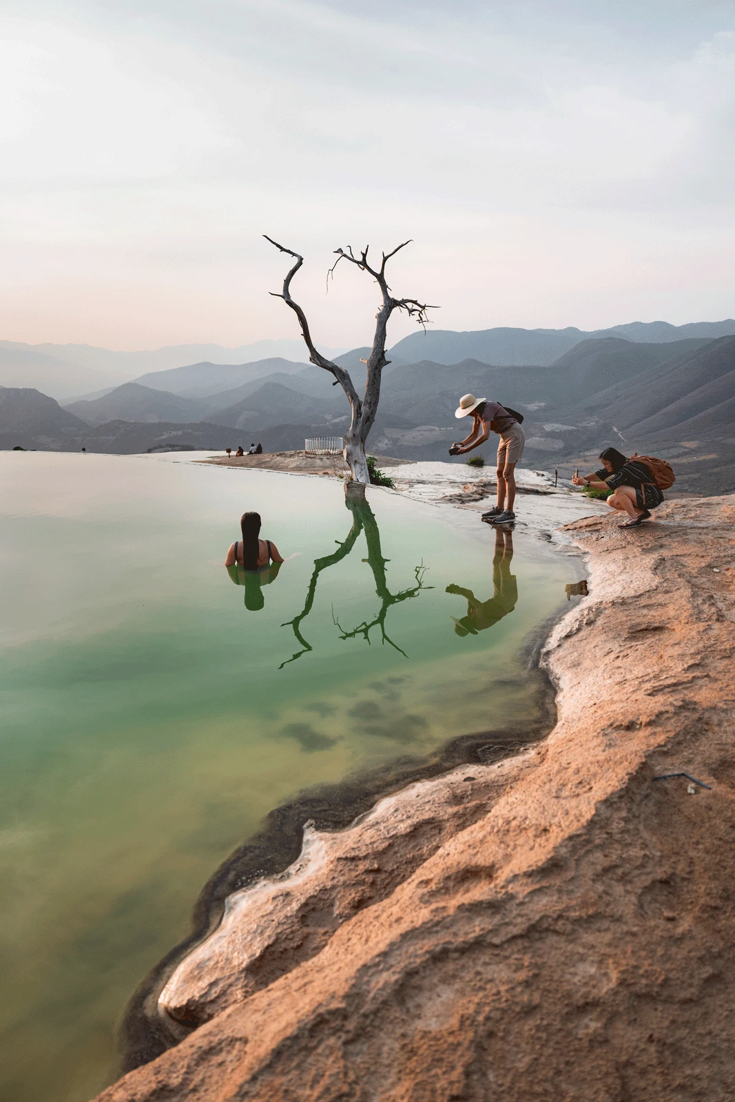 People enjoying the scenic view at Hierve el Agua in Mexico. One person is standing half-submerged in the water, while another person wearing a hat is leaning over to take a photo, with a barren tree and mountainous landscape in the background.