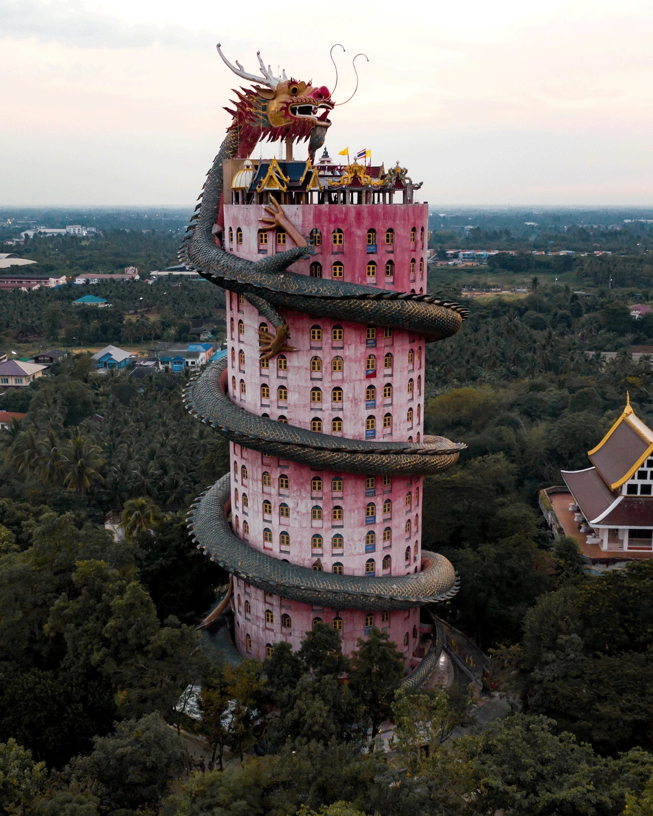 A tall pink cylindrical building with multiple floors stands amidst lush greenery. A massive intricate dragon sculpture coils around the building from the base to the top, where the dragon's head is prominent. The sky is overcast with a subtle glow from the setting sun.
