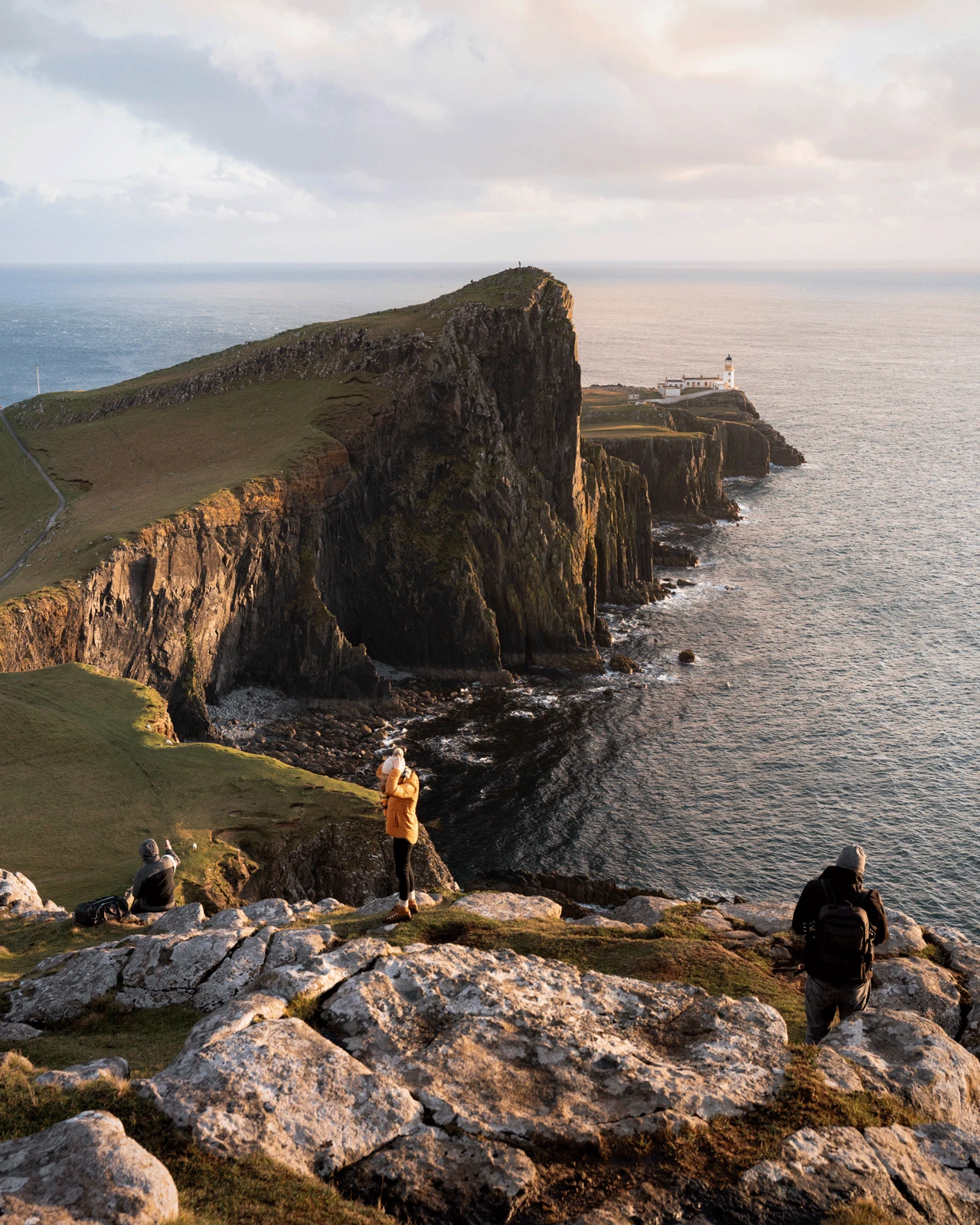 People are standing on a rocky cliff with a grassy terrain overlooking the ocean at sunset. A prominent cliff juts out into the sea with a lighthouse at the end. The sky has a soft, warm glow from the setting sun casting light on the landscape.