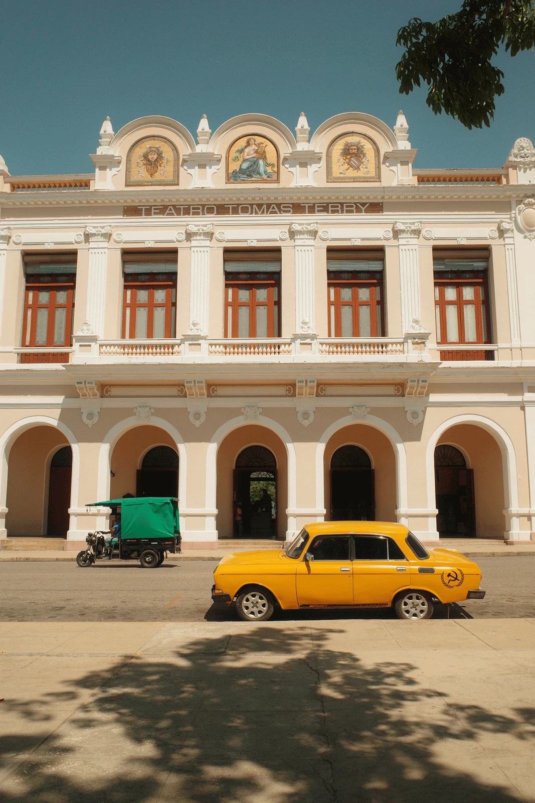 A yellow vintage car with a hammer and sickle symbol is parked in front of Teatro Tomás Terry. The building features three arches, grand columns, and ornate decorations, with three round paintings above. A green and black tuk-tuk is also parked nearby.