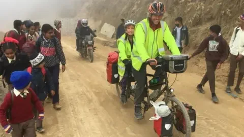 Tandem Travel Dave and Helen cycle on their Tandem on a mud road, surrounded by children and a motorbike behind them