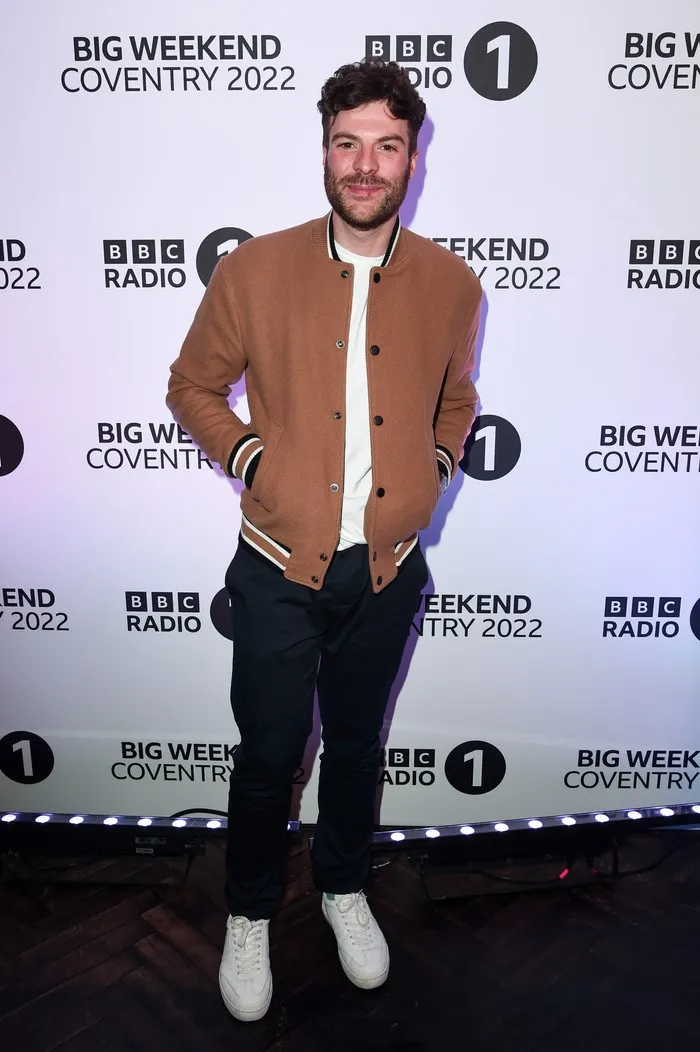 Jordan North posing in front of a white background for BBC Radio 1's Big Weekend. He is wearing a brown bomber jacket, a white t-shirt and black trousers.