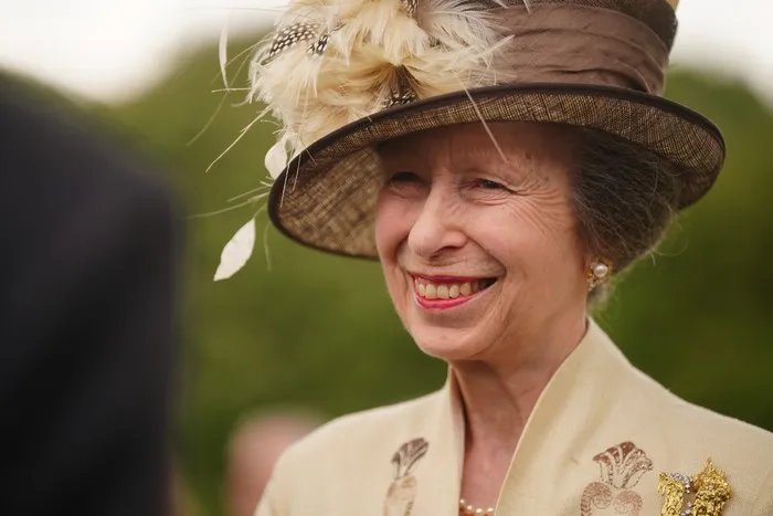 Princess Anne wearing a white suit and hat with a floral fascinator. She is smiling ahead.