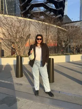 Woman standing outside wearing black sunglasses, black blazer, white t-shirt, blue straight leg jeans, shoulder bag